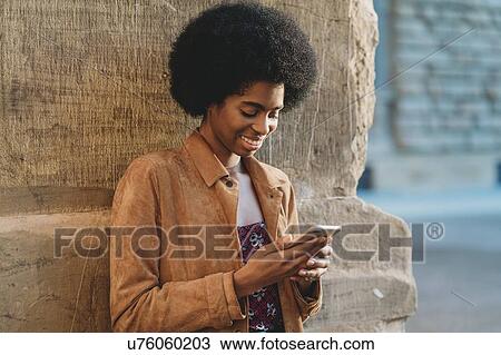 Stock Image - Young woman with afro hair using smartphone, leaning against stone wall. Fotosearch