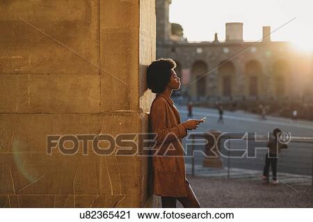 Stock Image - Young woman with afro hair using smartphone, leaning against stone wall, Florence, Toscana, Italy. Fotosearch
