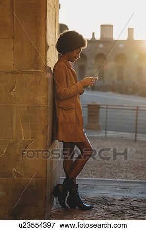 Stock Photo - Young woman with afro hair using smartphone, leaning against stone wall, Florence, Toscana, Italy. Fotosearch