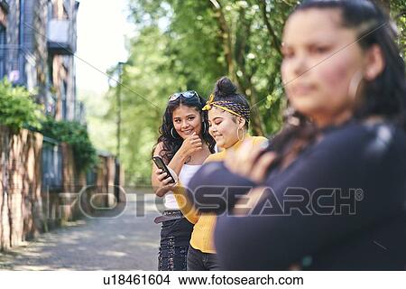 Picture - Young woman with teenage sisters looking at smartphone by park. Fotosearch