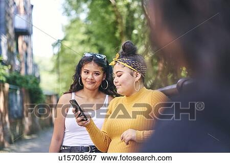 Stock Photo - Young woman with teenage sisters looking at smartphone by park, over shoulder view. Fotosearch