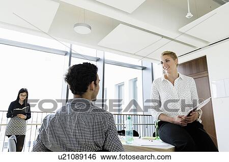Stock Photograph - Businessman and businesswoman talking in office, colleague in background. Fotosearch