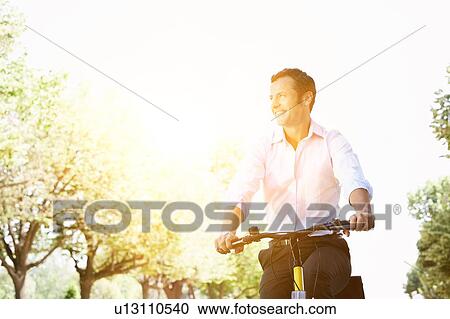 Stock Image - Businessman riding bicycle during sunny summer day in park. Fotosearch