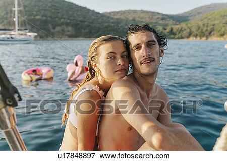 Stock Photo - Couple taking selfie, friends relaxing on floats in background, Italy. Fotosearch