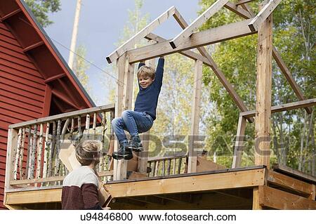 Stock Image - Father watching boy swinging on beam of unfinished treehouse. Fotosearch