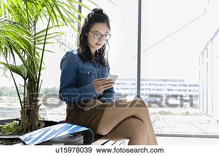 Stock Photo - Female student using smartphone beside glass wall in office building. Fotosearch