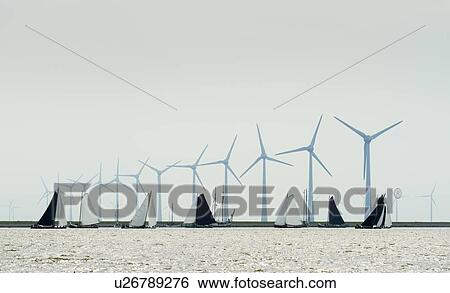 Stock Photograph - Flat-bottom freight ships participating in traditional 'Sktsjesilen' sailing race, large wind turbines in background, Lemmer, Friesland, Netherlands. Fotosearch