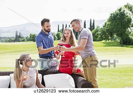 Stock Image - Friends enjoying wine and talking on sofa on golf course. Fotosearch