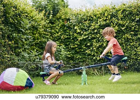 Stock Image - Girl and boy on toy seesaw in garden. Fotosearch