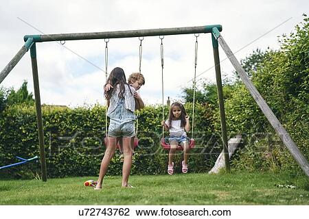 Stock Image - Girl carrying baby and watching younger sibling on swing. Fotosearch