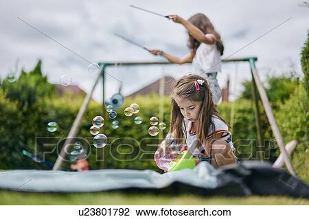 Stock Image - Girl playing with soap bubbles, sister in background with diabolo. Fotosearch