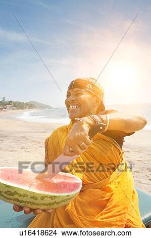 Picture - Happy mature Indian woman cutting watermelon at Vagator Beach. Fotosearch