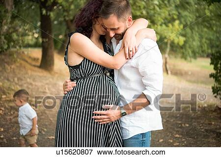 Stock Photo - Man hugging and touching belly of pregnant wife, son in background in park. Fotosearch