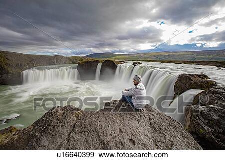 Stock Photo - Man sitting on edge of rock, Godafoss waterfall, Brardalur, Iceland. Fotosearch