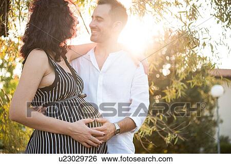 Stock Photograph - Man smiling and touching belly of pregnant wife in park. Fotosearch
