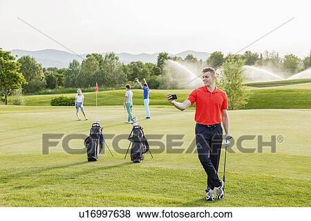 Man taking selfie on golf course, friends playing golf in background View Large Photo Image Stock Photo - Man taking selfie on golf course, friends playing golf in background. Fotosearch