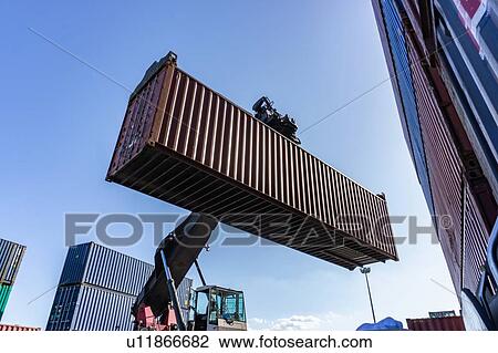 Stock Image - Reach stacker lifting shipping container in dock. Fotosearch