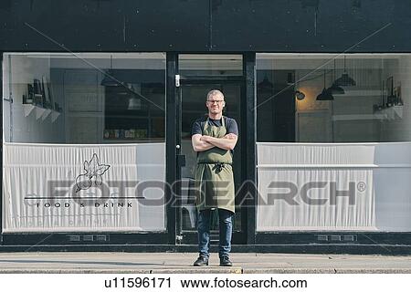 Stock Image - Restaurant owner with arms crossed at shopfront. Fotosearch