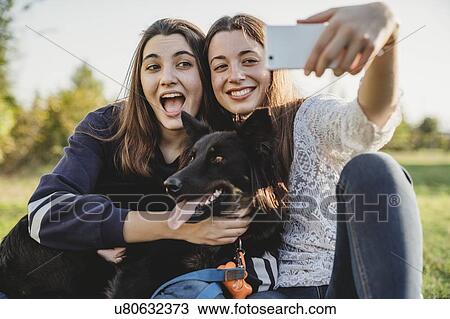 Stock Image - Sisters taking selfie with dog in park. Fotosearch