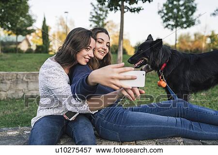 Stock Photo - Sisters taking selfie with dog in park. Fotosearch