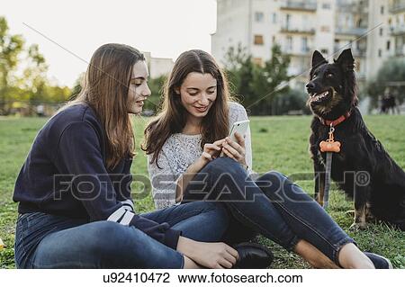 Stock Image - Sisters with dog talking in park. Fotosearch
