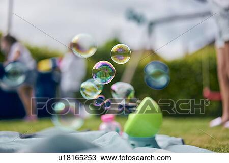 Stock Image - Soap bubble machine, family playing in garden in background. Fotosearch