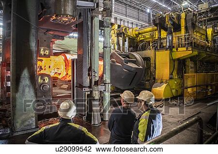 Stock Image - Steelworkers inspecting red hot steel ingot in heavy press in steelworks. Fotosearch