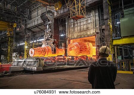 Stock Image - Steelworkers watching red hot steel ingots being craned out of furnace in steelworks. Fotosearch