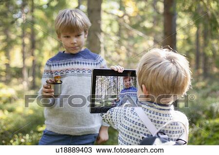 Stock Image - Toddler taking photograph of brother with cup of wild mushrooms in forest. Fotosearch
