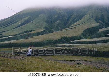 Stock Photograph - Woman sitting on ground admiring hillside, Landmannalaugar, Iceland. Fotosearch