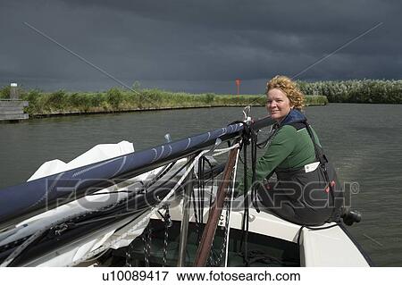Stock Photo - Woman steers sailing boat with lowered mast before crossing bridge, Friesland, Netherlands. Fotosearch