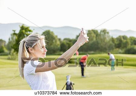 Picture - Woman taking selfie on golf course, friends playing golf in background. Fotosearch