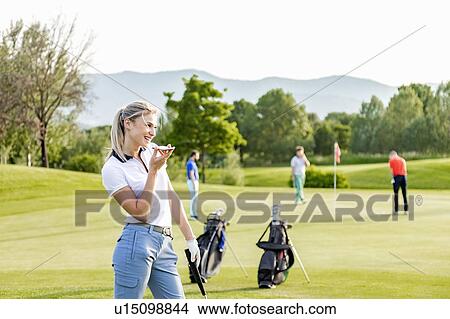 Picture - Woman talking on smartphone on golf course, friends playing golf in background. Fotosearch