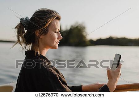 Stock Image - Woman using smartphone by lake, Bobcaygeon, Ontario, Canada. Fotosearch