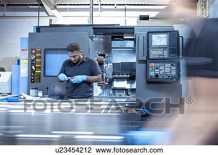 Stock Image - Worker inspecting machine parts on lathe in electronics assembly factory. Fotosearch