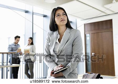 Stock Image - Young businesswoman in office, colleagues talking in background. Fotosearch