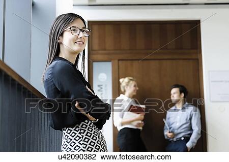 Stock Image - Young businesswoman in office, colleagues talking in background. Fotosearch