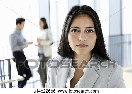 Stock Photograph - Young businesswoman in office, colleagues talking in background. Fotosearch