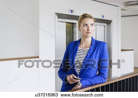 Stock Image - Young businesswoman leaning against railings in corridor of office building. Fotosearch
