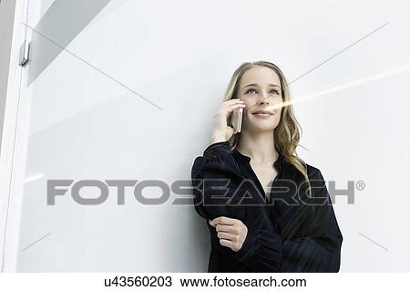 Stock Image - Young businesswoman talking on smartphone against white wall. Fotosearch