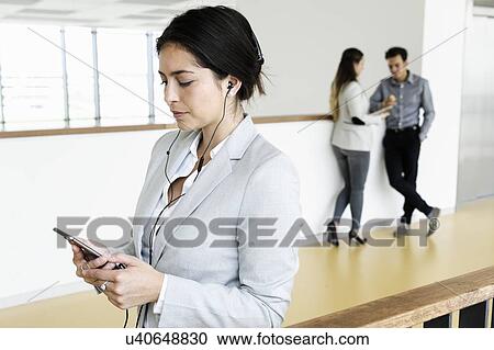 Stock Image - Young businesswoman using smartphone in office, colleagues talking in background. Fotosearch