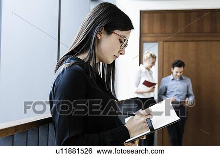 Young businesswoman writing notes in office, colleagues talking in background View Large Photo Image Stock Photograph - Young businesswoman writing notes in office, colleagues talking in background. Fotosearch