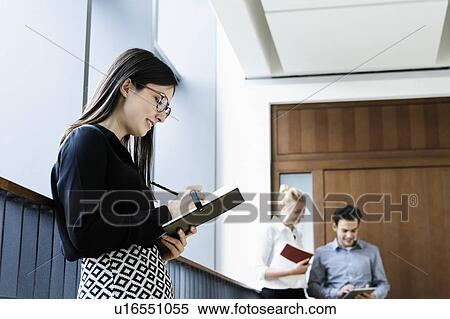 Stock Photography - Young businesswoman writing notes in office, colleagues talking in background. Fotosearch