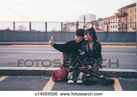 Stock Photograph - Young couple taking selfie on kerb, Milan, Italy. Fotosearch