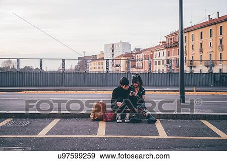 Stock Photo - Young couple texting on kerb, Milan, Italy. Fotosearch