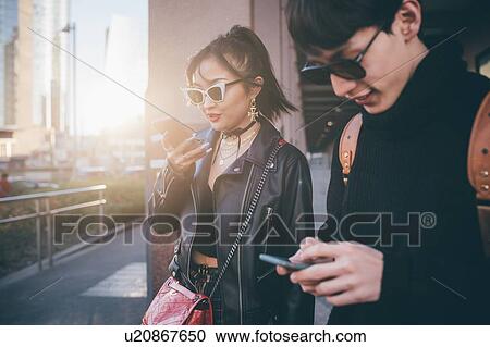 Stock Image - Young couple using mobile phone while walking on street, Milan, Italy. Fotosearch