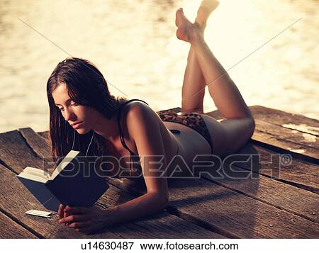 Stock Photo - Young woman in bikini lying on jetty reading book. Fotosearch