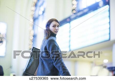 Stock Photography - Young woman in front of digital board in train terminal. Fotosearch