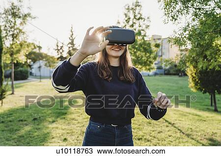 Stock Image - Young woman looking through VR headset in park. Fotosearch