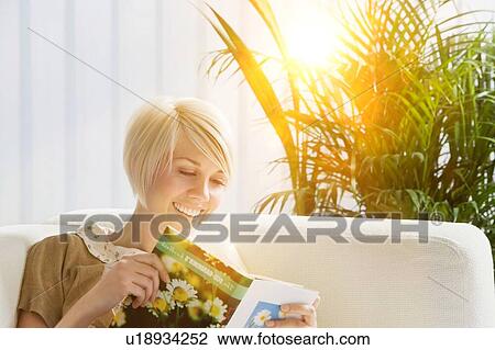 Stock Image - Young woman reading book on sofa. Fotosearch